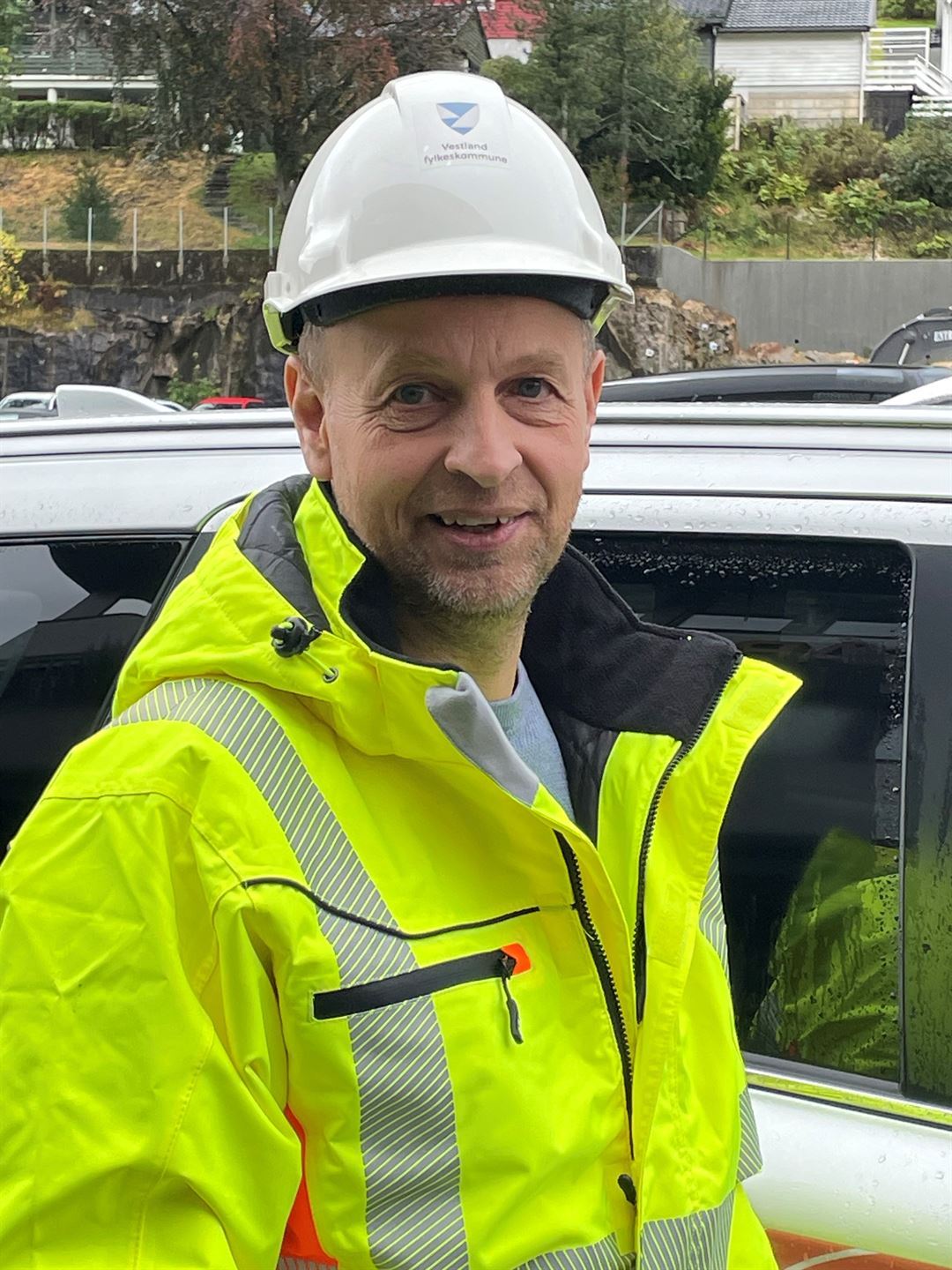 Mann med kvit hjelm og gul refleksjakke, står framfor ein bil.