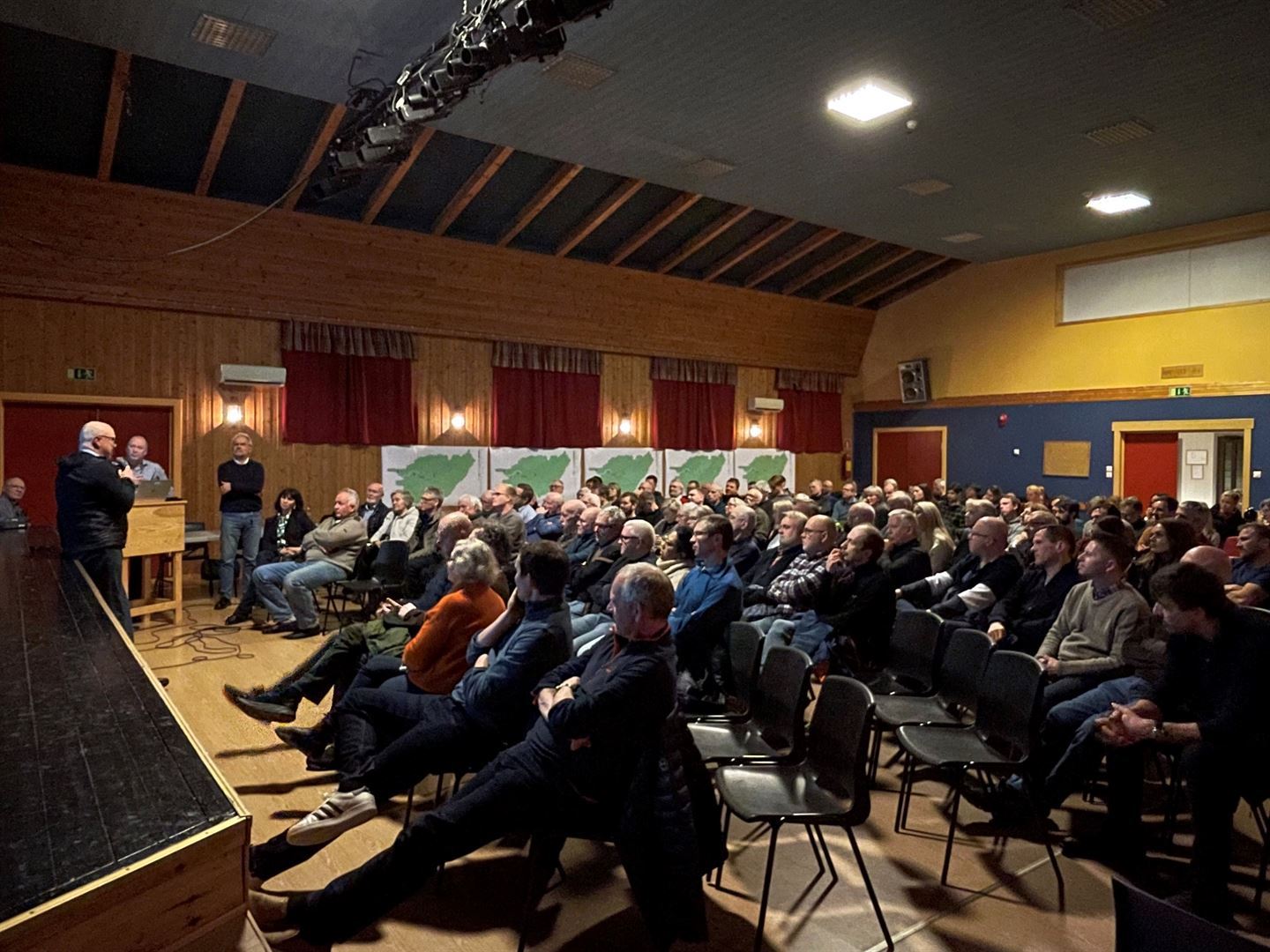 Mange folk sit på stolar i eit forsamlingslokale. Mann med mikrofon saman med to andre står oppreist.