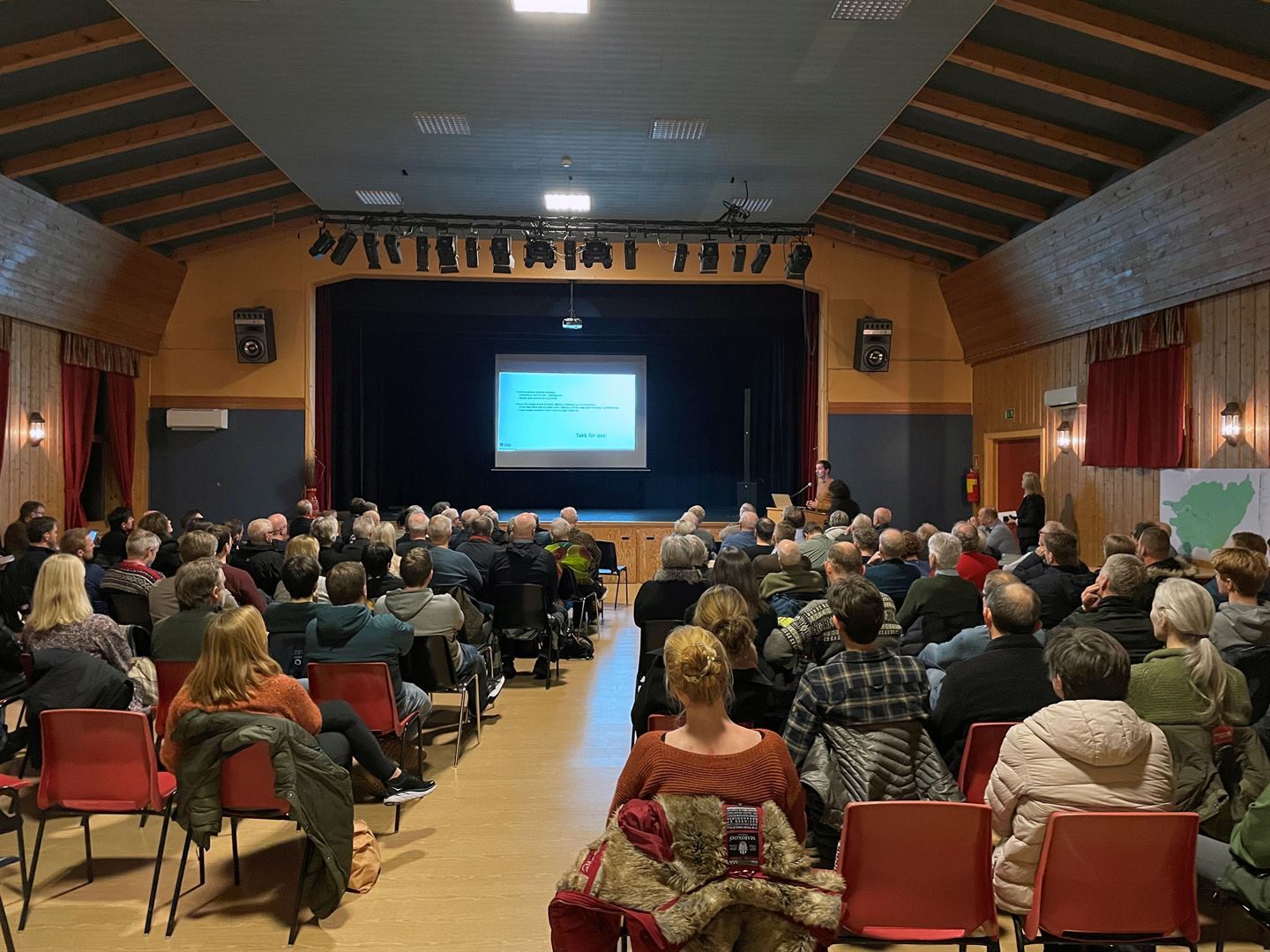 Mange menneske sit på rekke og rad i eit samfunnshus. Mann presenterer framme til høgre Lerret på scene viser ein powerpoint.