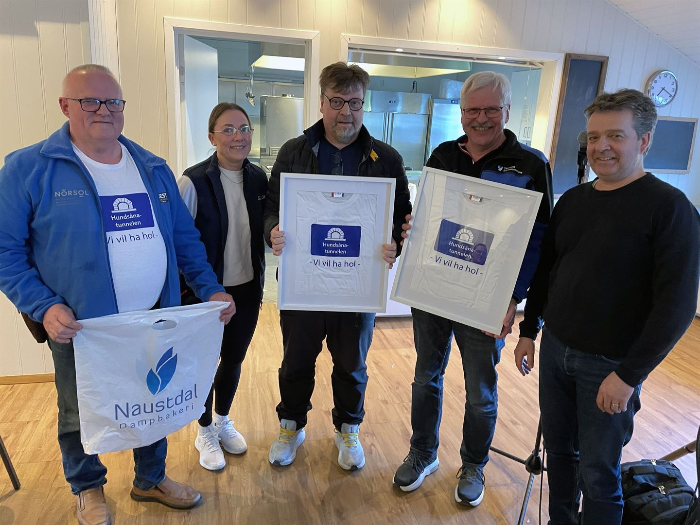 Fem personar står innandørs og held innramma T-skjorter og ein handlepose med tekst og logoar.