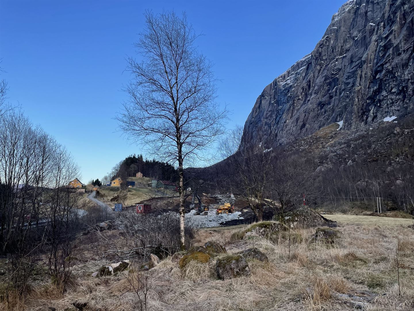 Landskap med grusveg, spreidde hus og tre i forgrunnen, og bratt fjellside til høgre under klar blå himmel.