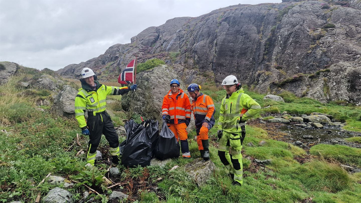 Fem menn med hjelm, nokon med gule og andre oransje arbeidskle med refleks. Står i eit kystlandskapap med fleire svarte søppelsekkar framfor seg. Den eine mannen held eit norsk flagg som har ødelagt flaggstang.
