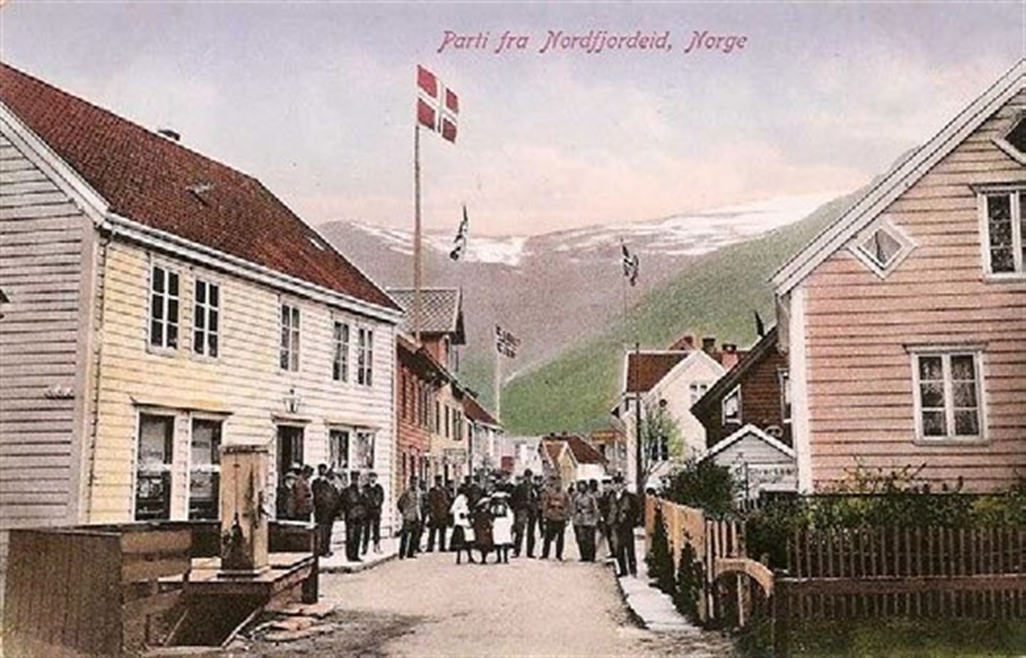 Postkort med bilete av Eidsgata som viser trehusbebyggjing med folk langs gata, og fleire hus som flaggar med norske flagg.