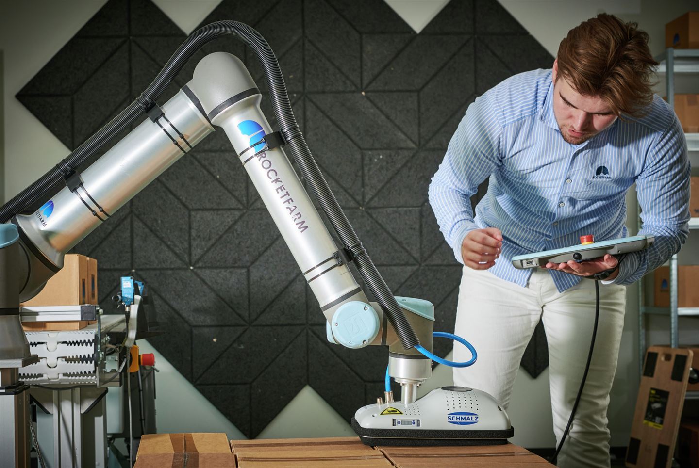 Ung mann i lyseblå skjorte, mørkt hår og skjegg jobbar med ein elektronisk penn på ein bærbar skjerm, gjennom dette styrer han ein stor robotarm i to ledd. På armen står det "Rocketfarm". Robotarmen set tape på pappesker. I bakgrunnen ser vi ein grå vegg med fliser og fleire ferdig tapa pappesker.